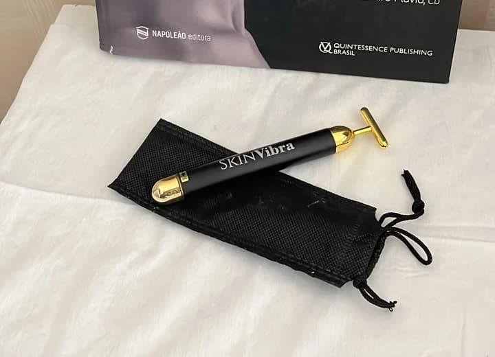 Massageador facial SkinVibra, bastão preto com detalhes dourados, sobre uma bolsa preta